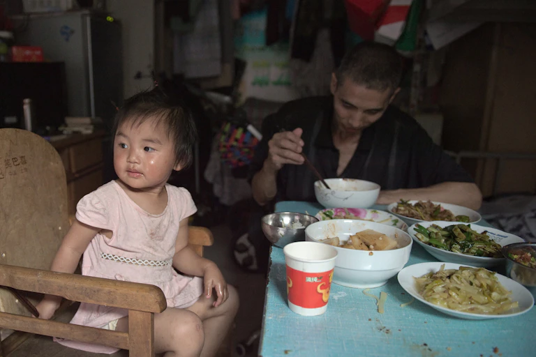 A Chinese toddler eats a big meal with her father in a cramped home.