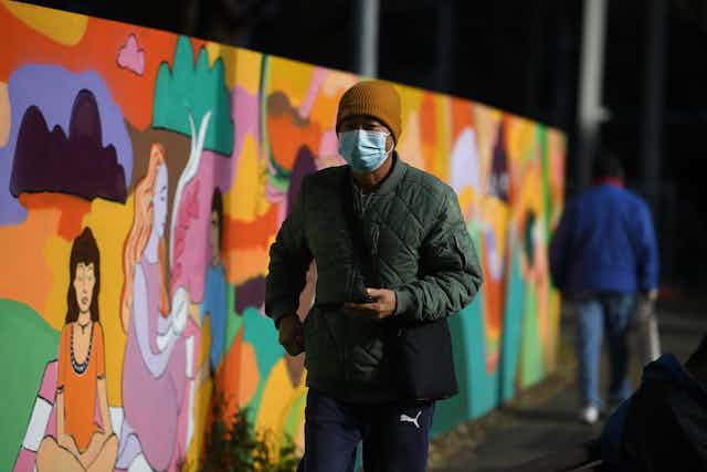 Man in mask walks next to colourful mural.