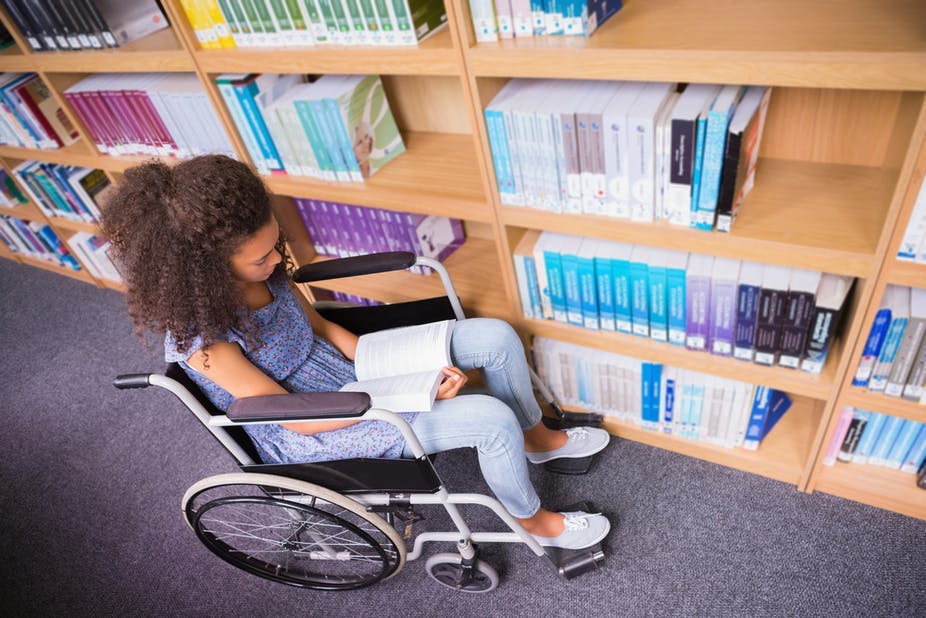 A young woman sitting on a wheelchair in a library and reading a book.