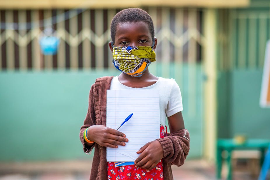 Child wit ha mask standing holding a notepad and a pen. 