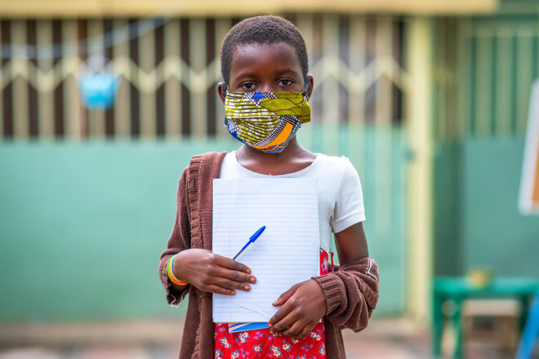 Child wit ha mask standing holding a notepad and a pen.
