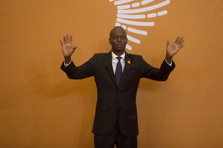 President Moïse in a black suit, raising his hands in front of an orange backdrop.