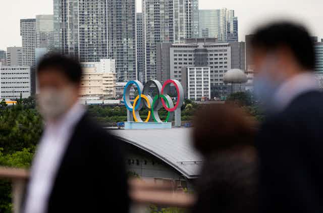 People in masks walk in the foreground as the Olympic rings are on a building in the background
