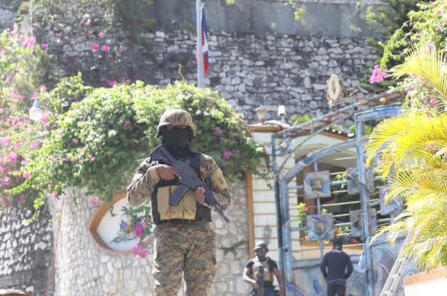 Heavily armed men in camp stand in a palm-dotted street; a Haitian flag is seen in the background.