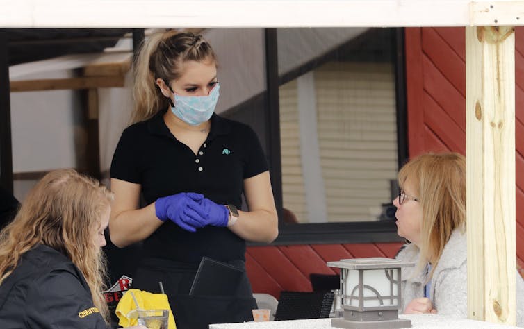 Masked and gloved waitress stands before two seated restaurant patrons