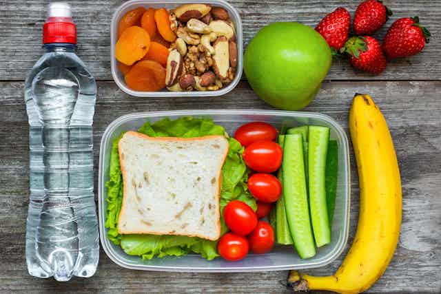 Lunchbox with cucumbers, tomatoes and a sandwich. A bottle of water and some fruit next to it and a separate lunch box with nuts and dried apricots.