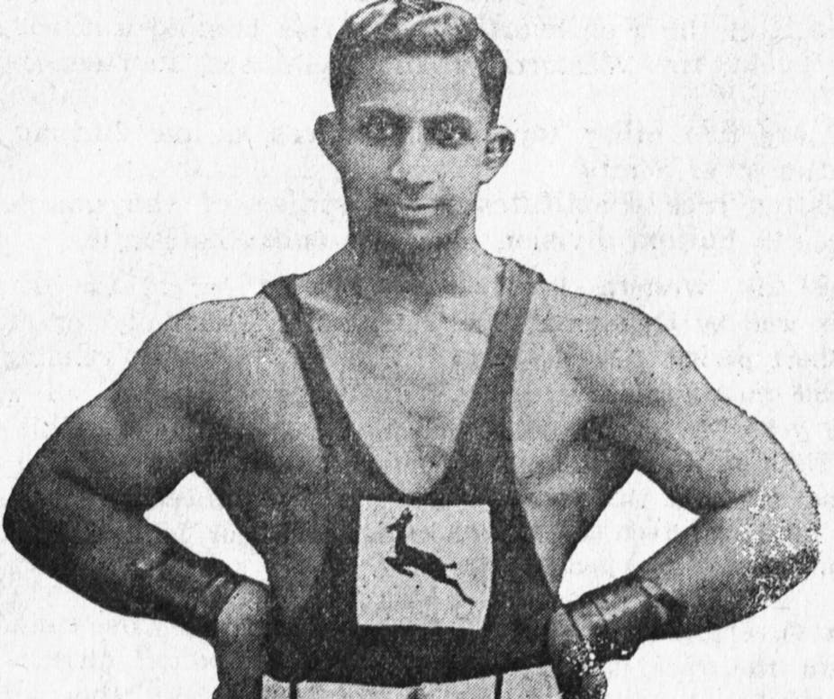Black and white photograph of well muscled man with arms akimbo, wearing wrist supports and athletic vest with Springbok emblem