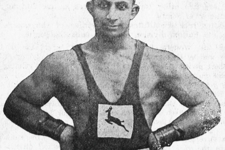 Black and white photograph of well muscled man with arms akimbo, wearing wrist supports and athletic vest with Springbok emblem