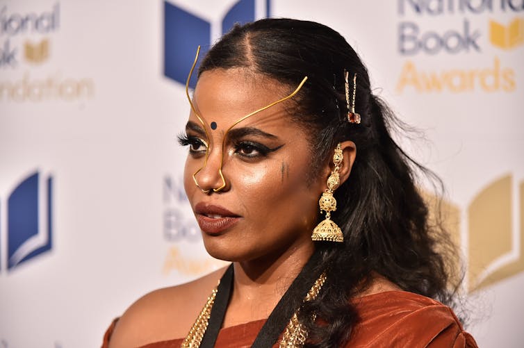 A young person presenting as a woman against a backdrop for a book event, thin gold face jewellery distinguishes her look.