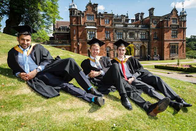 Three male students in graduation gowns and caps recline on a lawn outside a university building