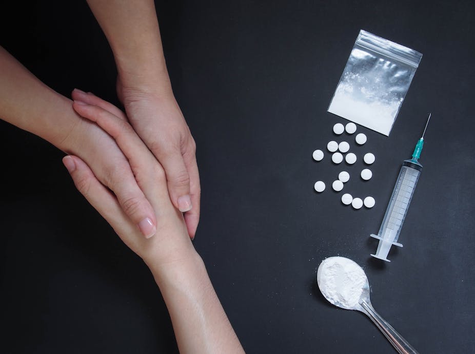 A person holding another person's hand supportively, with illicit drugs on a table beside them, to illustrate support and recovery.