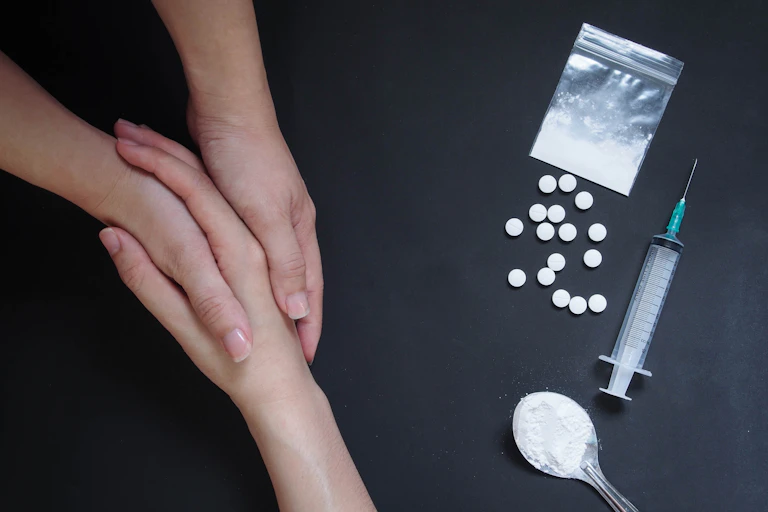 A person holding another person's hand supportively, with illicit drugs on a table beside them, to illustrate support and recovery.