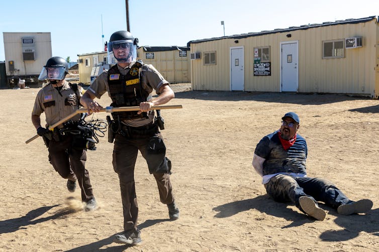 Two police in riot gear run while handcuffed man sits on ground