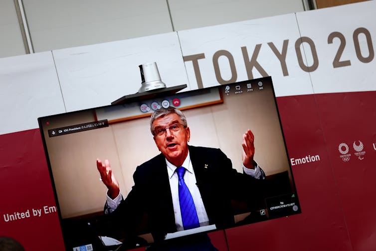 Bach raises his hands while appearing on a TV screen at a news conference in Tokyo.