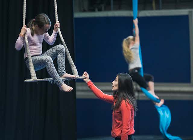 A girl on a trapeze receiving instruction, with another girl climbing a rope in the background