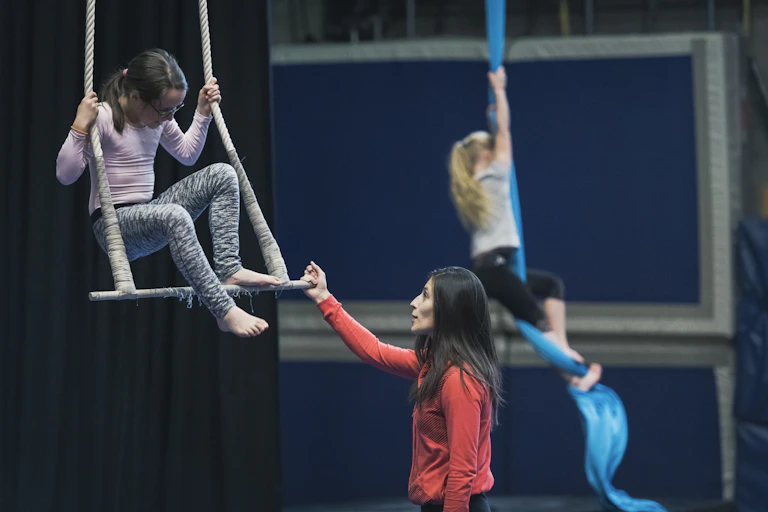 A girl on a trapeze receiving instruction, with another girl climbing a rope in the background