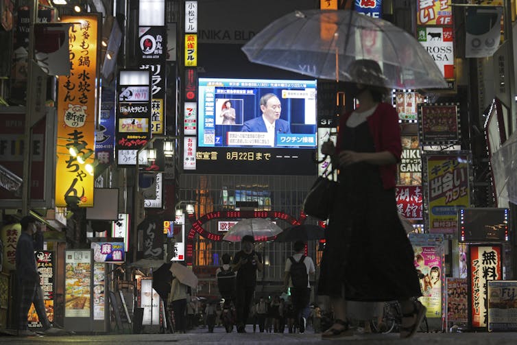 A night time image of people on a rainy street with a large screen in the background.