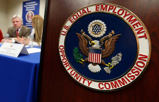 The U.S. Equal Employment Opportunity Commission seal is displayed in the foreground as a man and a woman chat at a table with a blue cloth draped over it. 