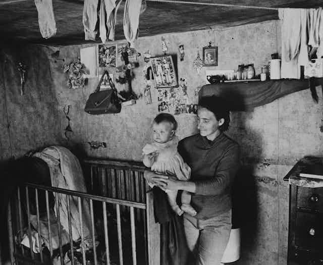 A mother holds her child in a room with a crib and lots of little pictures and other things hanging from the wall in a black and white photo in Appalachia.