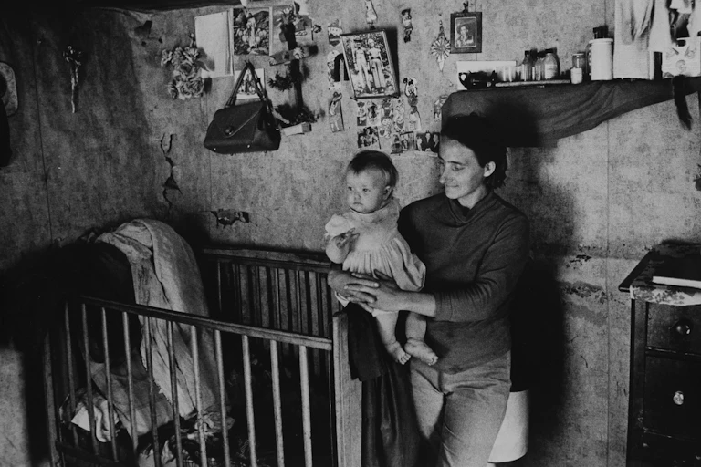 A mother holds her child in a room with a crib and lots of little pictures and other things hanging from the wall in a black and white photo in Appalachia.