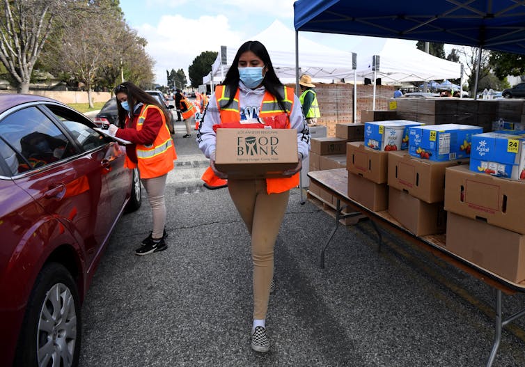 A woman carries a box of food to a car waiting at a food bank in Los Angeles