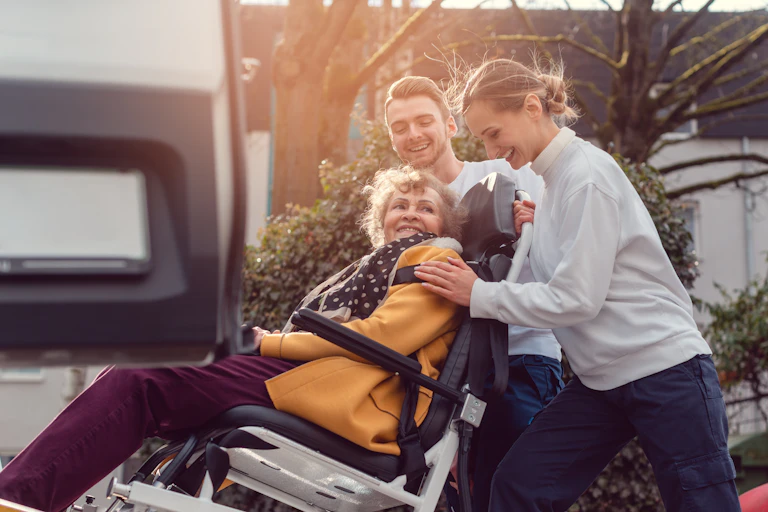 Two young people assist a woman with a disability into a vehicle.