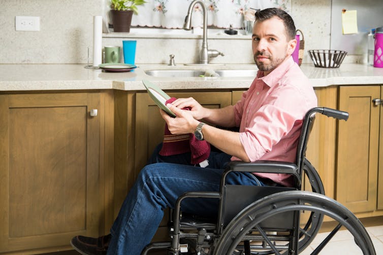 A man in a wheelchair drying a plate in the kitchen.
