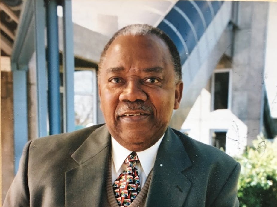 A portrait of an elder man in a suit and tie, a slight smile on his lips as he looks into camera; behind him angular architectural features.