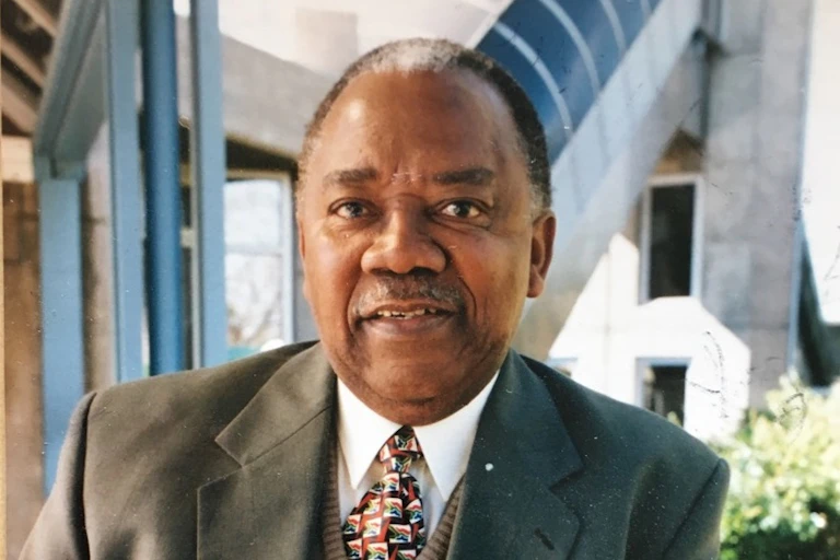 A portrait of an elder man in a suit and tie, a slight smile on his lips as he looks into camera; behind him angular architectural features.