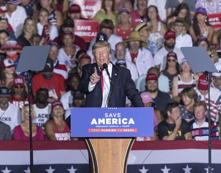 Former US President Donald Trump speaking at a rally. There is a Save America placard on his podium, and he has one finger raised.