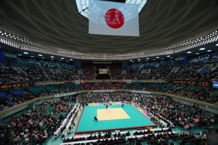 Seated crowds in an arena watch a judo match with Japanese flag above