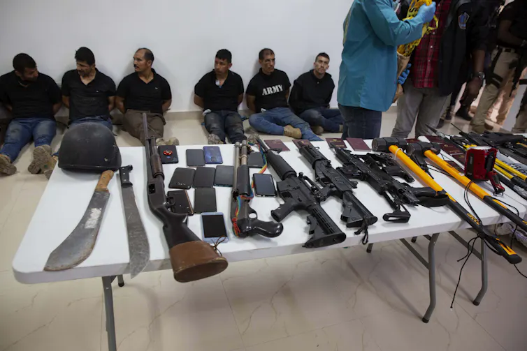 Weapons on a table, including machetes and assault rifles. Behind them, a row of men sit.