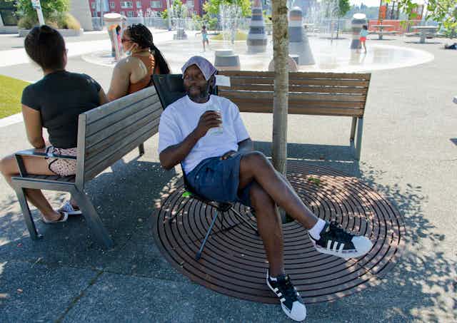 A Black man in shorts and a T-shirt sits under a tree. He is drinking water and has a cloth on his head. In the background, children play in a spray park.