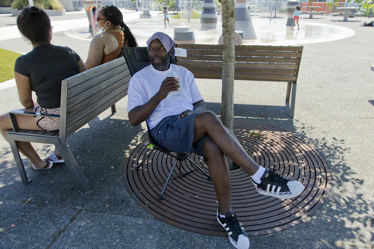 A Black man in shorts and a T-shirt sits under a tree. He is drinking water and has a cloth on his head. In the background, children play in a spray park.