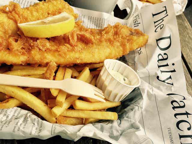 Fried cod on a bed of chips with a slice of lemon and tartare sauce wrapped in newspaper.