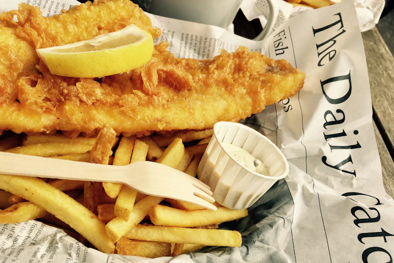 Fried cod on a bed of chips with a slice of lemon and tartare sauce wrapped in newspaper.