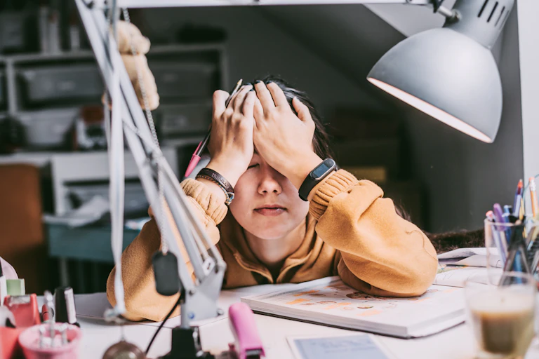 A student rests their head against their hands on a cluttered desk.