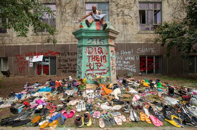 Spray paint reads 'dig them up' '215' and 'shame' with a bunch of shoes in the foreground