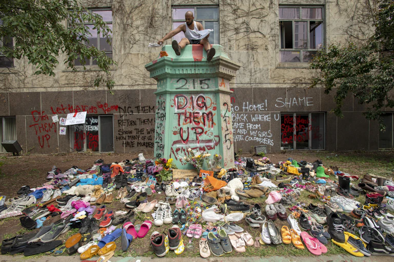 Spray paint reads 'dig them up' '215' and 'shame' with a bunch of shoes in the foreground