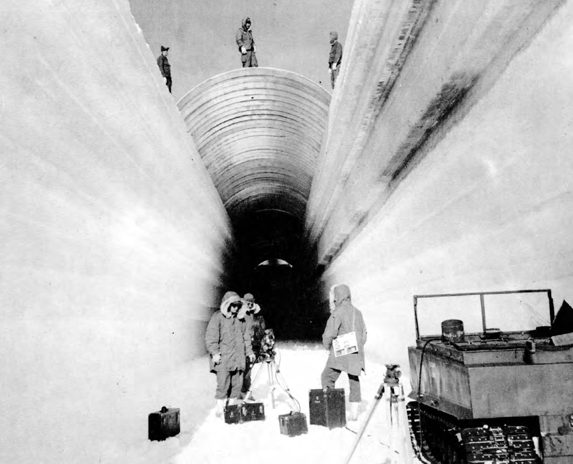 Three people stand at the opening of an icy trench with a half-round metal cover