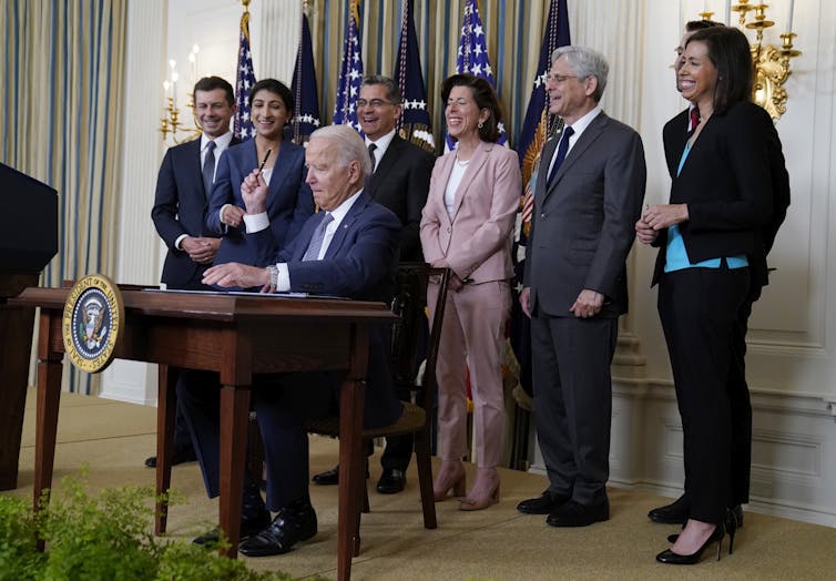 President Joe Biden sits at a desk as he holds a pen in his right hand and hands it to one of the government officials standing behind him.