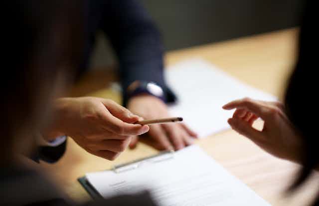 Two people are sitting at a table with a business contract on it as the right hand of the person on the opposite side hands a pen across the table.