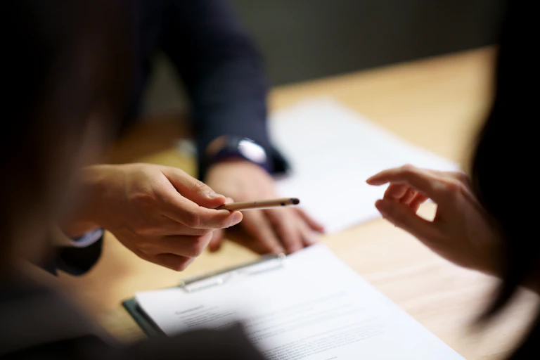 Two people are sitting at a table with a business contract on it as the right hand of the person on the opposite side hands a pen across the table.