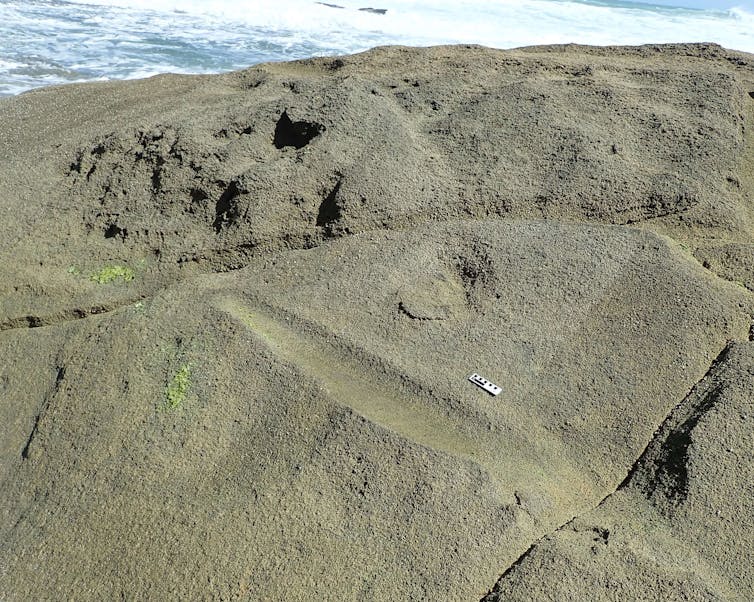 A smooth grey rock surface, with some pock marks, alongside the blue ocean. There is a mark in the rock's surface that looks like a deep, diagonal groove.