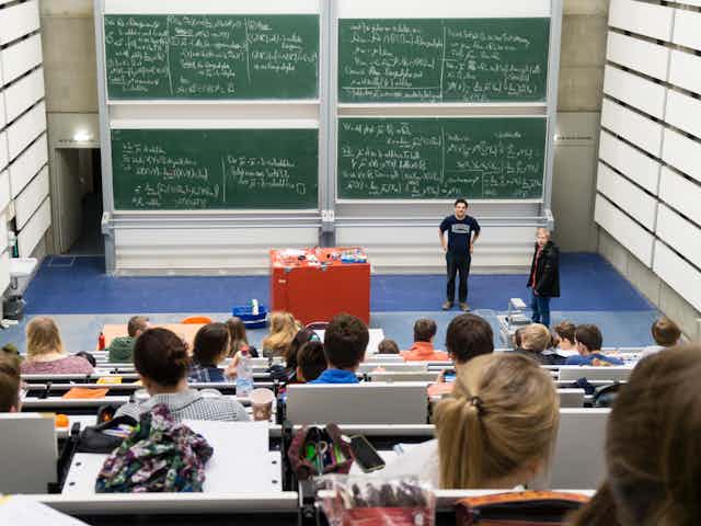 Students in an auditorium look down at professors and a wall of blackboards covered in mathematical formulae