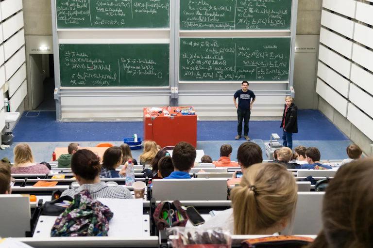 Students in an auditorium look down at professors and a wall of blackboards covered in mathematical formulae
