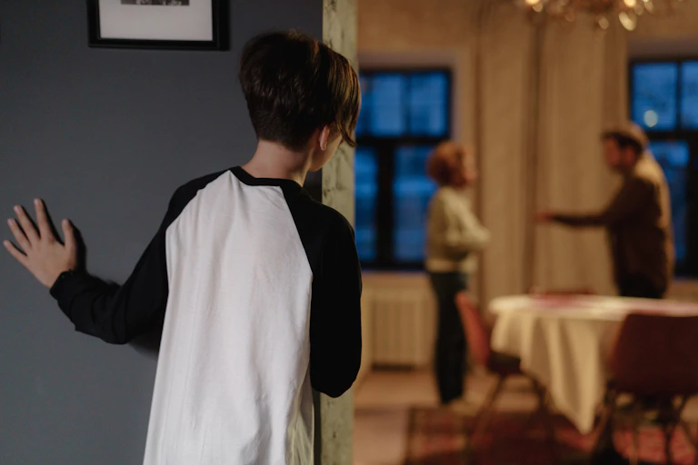 Boy peeks around corner as parents argue in the background