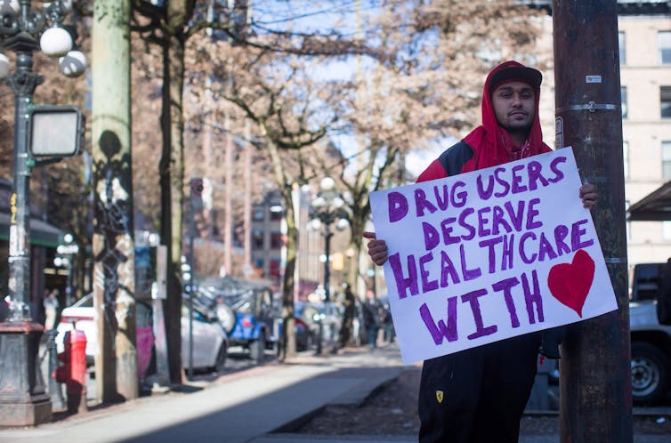 A man holds a sign that reads Drug Users Deserve Health Care with Heart.