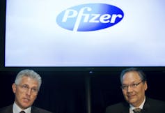 Two men sit in front of a Pfizer sign.
