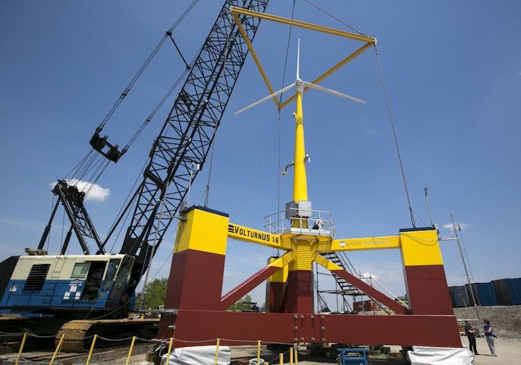 People stand next to a small wind turbine held by a crane. Just the base is three times higher than a human.